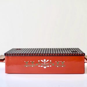 Op de afbeelding: Een rode metalen tafelgrill met een zwart grillrooster en houten handvatten. De grill heeft een wit en groen geometrisch ontwerp.