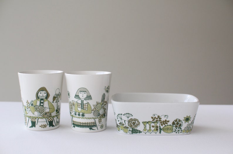 Op de afbeelding: Set van drie witte keramische bekers en een rechthoekige schaal met een groen en bruin bloemenpatroon. De bekers hebben illustraties van mensen in traditionele kleding.