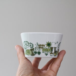 Op de afbeelding: Een witte keramische kom met een groen en bruin bloemenpatroon. De kom wordt in een hand gehouden.