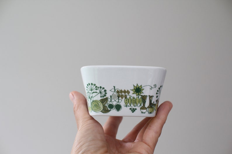 Op de afbeelding: Een witte keramische kom met een groen en bruin bloemenpatroon. De kom wordt in een hand gehouden.