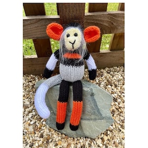 May include: A knitted monkey toy with black, white, and orange stripes. The monkey has a long, white tail and is sitting on a gray rock.