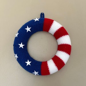 May include: A knitted wreath in the colors of the American flag. The wreath is blue with white stars on one side and red and white stripes on the other side.