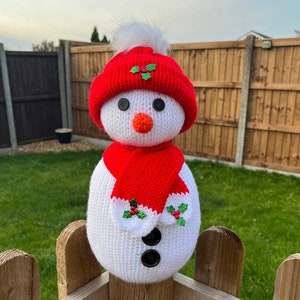 May include: A white knitted snowman wearing a red knitted hat with a green holly design and a red knitted scarf with green holly accents. The snowman is standing on a wooden fence.