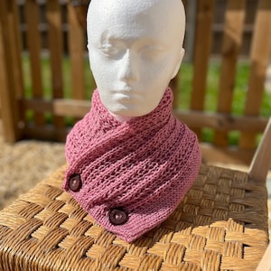 Könnte beinhalten: Ein rosafarbener gestrickter Schal mit Stehkragen und zwei braunen Knöpfen. Der Schal wird auf einem Korbstuhl präsentiert.