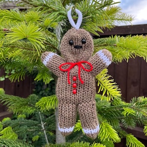 May include: A knitted gingerbread man ornament with white trim, red buttons, and a red bow. The ornament is hanging from a Christmas tree.