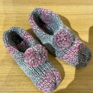May include: A pair of grey and pink knitted slippers with fluffy pink pom-poms on the top.