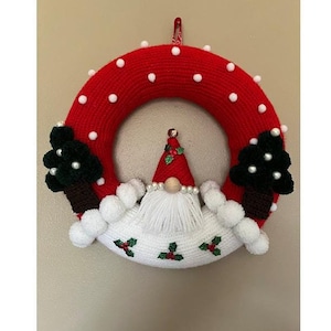 Pode incluir: Uma guirlanda de Natal vermelha e branca de crochê com um gnomo no centro. O gnomo usa um chapéu vermelho com azevinho e uma barba branca. A guirlanda é decorada com pompons brancos e azevinho verde.