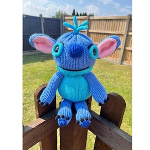 Może przedstawiać: Pluszowa zabawka w kolorze niebieskim i turkusowym przypominająca Stitch z filmu Disneya Lilo & Stitch. Zabawka ma duże oczy, różowe uszy i czarne łapy.