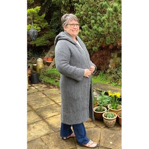 Puede incluir: Un cárdigan gris con capucha de corte largo y holgado. El cárdigan está tejido y tiene un patrón texturizado. La persona que lleva el cárdigan está de pie en un jardín.
