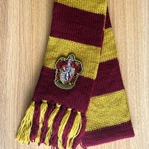 Pode incluir: Cachecol de malha com listras bordô e douradas. Apresenta um patch com o brasão da Grifinória e franjas douradas. Feito de um material macio e quente, ideal para o inverno.