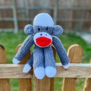 May include: A gray knitted sock monkey with a white face and red lips sitting on a wooden fence.
