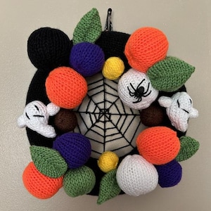 Pode incluir: Uma grinalda de Halloween feita à mão em forma de cabeça de Mickey Mouse preta. É adornada com elementos de malha laranja, roxa, amarela e verde, incluindo abóboras, folhas e formas redondas. Fantasmas de malha brancos e uma teia de aranha completam o design festivo.