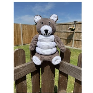 Può includere: Un orsacchiotto fatto a mano in maglia marrone e bianco, con naso nero e dettagli bianchi. L'orso è seduto su una staccionata di legno, con cielo blu ed erba verde sullo sfondo. L'orso ha un design unico a corpo impilato.