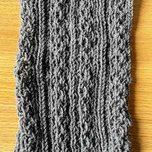 Könnte beinhalten: Nahaufnahme einer grauen Strickmütze mit einem strukturierten Muster. Die Mütze ist aus weichem, warmem Garn gefertigt und hat ein schlichtes, elegantes Design.