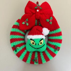 May include: A green and red striped knitted Christmas wreath with a Grinch face and a red bow with holly accents.