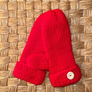 May include: Vibrant red knitted mittens with a turned-up cuff. A small, round wooden button with the words "handmade with love" is sewn on the cuff. The mittens are made of a thick, textured yarn.