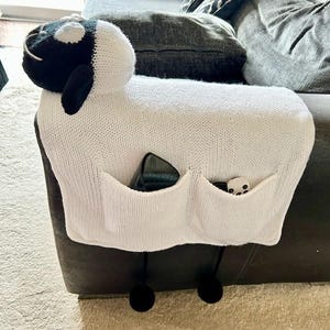 May include: A white knitted sheep-shaped couch armrest cover with two pockets. The sheep has a black and white head and black pom-poms hanging from the bottom. A remote control and a small toy are in the pockets.