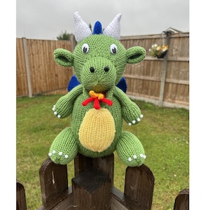 May include: A handmade green knitted dragon toy with white horns, blue wings, and a yellow belly. The dragon has white accents on its feet and a red and yellow collar. It is sitting on a wooden fence.