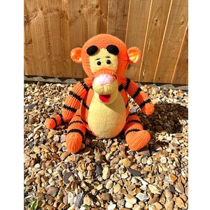 May include: A crocheted orange and black striped tiger plush toy with a yellow face and red tongue. The toy is sitting on a bed of small brown stones.