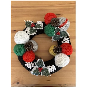 May include: A handmade Christmas wreath with a black base, adorned with red, green, white, and gray yarn balls. The wreath is decorated with holly leaves, white berries, and pinecones, creating a festive holiday decoration.