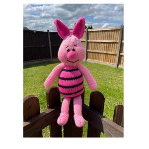 May include: A pink and black striped knitted pig toy with a smiling face. The toy is sitting on a wooden fence.