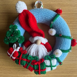 May include: A blue knitted Christmas wreath with a gnome wearing a red hat and white beard. The wreath is decorated with green and red knitted presents, a small knitted Christmas tree, and white pom-poms.