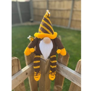 Könnte beinhalten: Ein brauner und gelber gestrickter Zwerg mit einer gestreiften Mütze und passenden gestreiften Beinen. Der Zwerg sitzt auf einem Holzzaun mit zwei kleinen gelben und schwarzen Bienen auf seiner Mütze und seinem Arm.