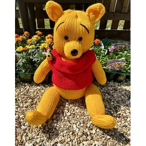 May include: A knitted Winnie the Pooh bear wearing a red shirt and sitting on a bed of small stones. A small knitted bee is attached to the bear's shirt.