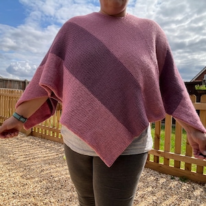 May include: A pink and purple knitted poncho with a diagonal stripe pattern. The poncho is worn by a person standing in a backyard.