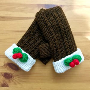 Könnte beinhalten: Braune gestrickte Fingerlose Handschuhe mit weißen und grünen gehäkelten Manschetten und roten Stechpalmen-Beerenverzierungen.