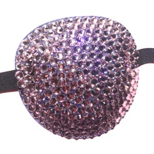 May include: A purple eye patch adorned with numerous small, sparkling rhinestones. The oval-shaped patch is attached to a black elastic band. The rhinestones are in shades of purple and pink.