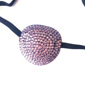 May include: An eye patch featuring a circular front panel densely covered in small, shimmering, light purple rhinestones. The patch is attached to a thin, plain black elastic strap that extends from the sides to secure the item in place.