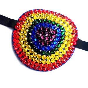 May include: An oval eye patch featuring concentric circles of faceted rhinestones in a rainbow colour sequence, starting with a dark purple centre, followed by rings of blue, green, yellow, and red, attached to a black elastic strap.