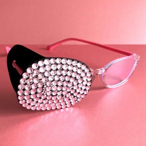 May include: Cat-eye sunglasses with clear frames, pink lenses and arms. One lens is covered with a black velvet patch, densely encrusted with clear rhinestones. The sunglasses rest on a pink surface.