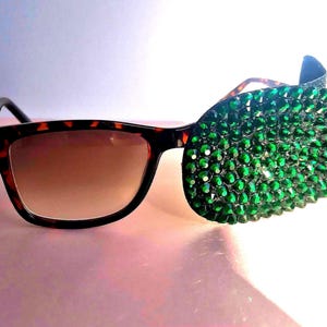 May include: Sunglasses with a tortoiseshell frame and gradient lenses. The right lens is covered in green rhinestones. The sunglasses have a classic shape with a modern, embellished twist, offering a unique and eye-catching design.