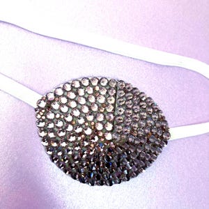 May include: An oval eye patch covered in shimmering, multi-tonal purple and dark violet rhinestones. The patch features a gradient effect, transitioning from lighter, reflective crystals to deeper, darker shades, and is attached to a white elastic strap.