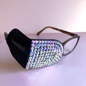 May include: A black eye patch, embellished with iridescent rhinestones, is attached to a pair of spectacles with a tortoiseshell frame. The patch covers the left lens, providing a decorative and functional accessory.