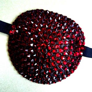 May include: A circular eye patch covered in numerous small, faceted, deep red crystals. The patch is attached to a thin, black elastic strap that extends from both sides of the circular piece, designed to secure the item in place.