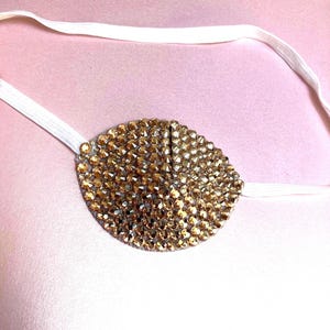 May include: An oval-shaped eye patch covered in shimmering gold-toned rhinestones. The patch features a white elastic strap attached to both sides, designed to secure the piece comfortably in place while providing a decorative, reflective finish.