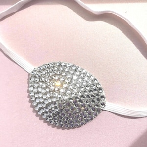 May include: A white eye patch with a silver rhinestone design. The eye patch is attached to a white elastic band.