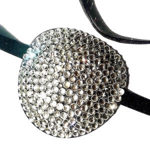 May include: A black headband with a round, silver rhinestone embellishment. The rhinestones are arranged in a gradient pattern, with larger rhinestones in the centre and smaller rhinestones around the edges.