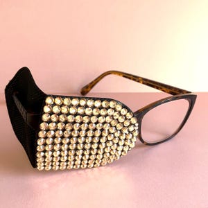 May include: Spectacles featuring a tortoiseshell frame and a black side shield attachment on the left lens. The side shield is covered in numerous small, clear, faceted crystals that reflect light, creating a sparkling, textured appearance.