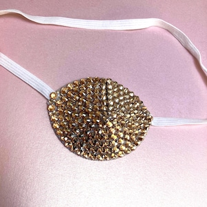 May include: An oval-shaped eye patch covered in small, reflective gold-toned rhinestones. The patch features a vertical seam down the centre and is attached to a thin, white elastic strap designed to secure the piece around the head.