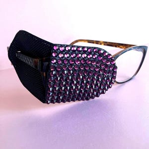 May include: An eye patch for glasses, with a black fabric backing and a surface covered in small, sparkling purple rhinestones. The patch is attached to a pair of spectacles with tortoiseshell arms.