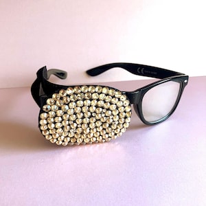 May include: Black-framed glasses with a decorative eye patch. The eye patch is oval-shaped and covered in small, sparkling, gold-coloured rhinestones. The glasses have clear lenses against a light pink background.