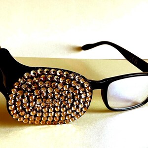 May include: Black-framed reading glasses with a decorative eye patch. The eye patch is black and covered in small, sparkling, gold-coloured rhinestones. The glasses have a rectangular lens shape.