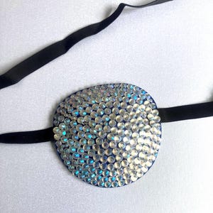 May include: An oval eye patch featuring a dense arrangement of clear, iridescent rhinestones on a blue base. The eye patch is attached to a thin, black elastic strap designed to secure the piece in place, providing a functional and decorative accessory.