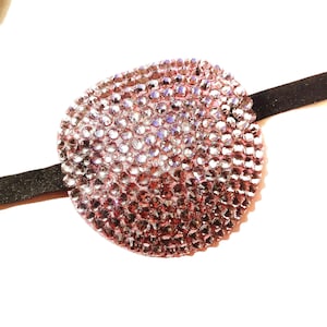 May include: A circular eye patch covered in shimmering light pink and silver rhinestones. The patch is attached to a thin, plain black elastic strap that extends horizontally from both sides of the circular, crystal-encrusted centrepiece.