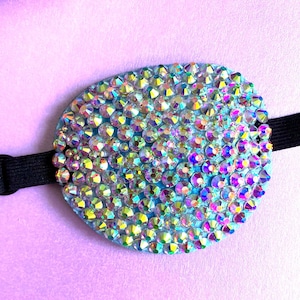 May include: An oval eye patch featuring a light blue base densely covered in iridescent, multi-faceted rhinestones that reflect various colours. The patch is attached to a black elastic strap extending from both sides of the oval shape.