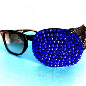 May include: A pair of sunglasses with a blue rhinestone eye patch covering one lens. The sunglasses have a brown frame and dark lenses.